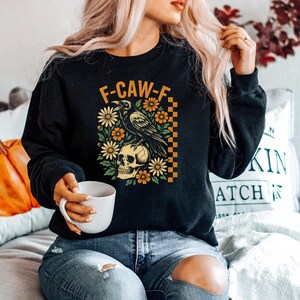 May include: Black sweatshirt with a graphic featuring a raven perched on a skull, surrounded by flowers. The text "F-CAW-F" is above the design. The design has orange, yellow, and green accents. The sweatshirt is a crew neck style.