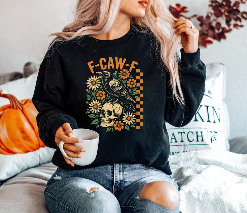May include: Black sweatshirt with a graphic featuring a raven perched on a skull, surrounded by flowers. The text "F-CAW-F" is above the design. The design has orange, yellow, and green accents. The sweatshirt is a crew neck style.