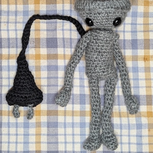 May include: A handmade gray crochet alien doll with black button eyes and a black cord attached to a black cone-shaped object. The doll is on a checkered fabric background.