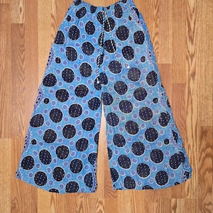 May include: Wide-leg pants featuring a blue background with a repeating pattern of large black circles containing white dashed lines. The pants have an elasticized waistband with a black and white braided drawstring cord and side pockets.