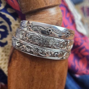 May include: Three silver bangles with intricate floral designs are stacked on a wooden form. The bangles have detailed patterns of flowers and leaves, with a polished finish. The background includes colorful fabric.