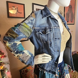 Könnte beinhalten: Blaue Jeansjacke mit Patchwork-Ärmeln. Die Ärmel sind mit verschiedenen Stoffflicken in Blau-, Grün- und Grautönen verziert. Die Jacke hat einen klassischen Kragen, Knopfverschlüsse und zwei Brusttaschen. Die Schaufensterpuppe trägt ein beiges Häkeltop.