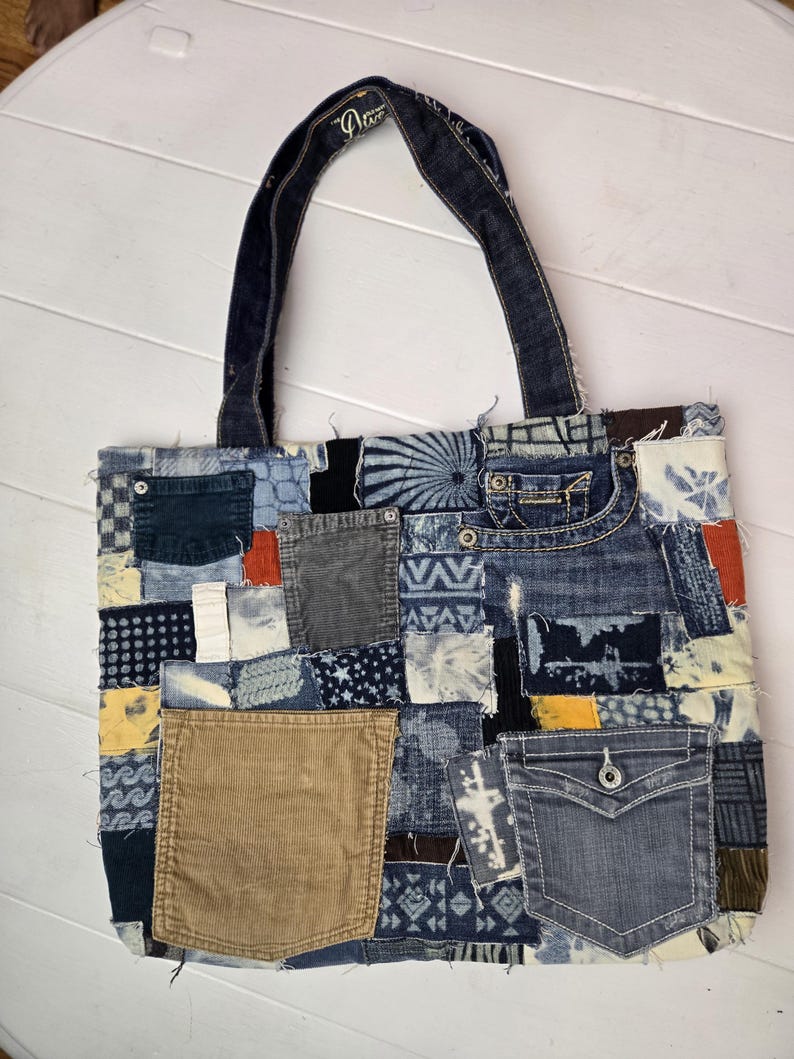 May include: A patchwork denim tote bag with a variety of blue denim patches, corduroy, and other fabrics. The bag features a denim handle and a pocket on the front. The bag is a unique, handmade item.