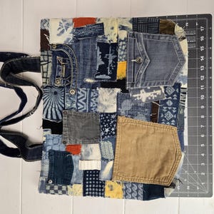 May include: A patchwork tote bag made from repurposed denim. The bag features a variety of denim washes and textures, including pockets and decorative stitching. The bag has dark blue denim handles.