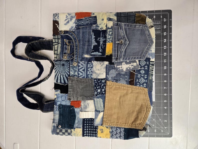 May include: A patchwork tote bag made from repurposed denim. The bag features a variety of denim washes and textures, including pockets and decorative stitching. The bag has dark blue denim handles.