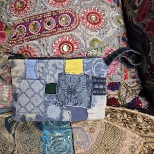 Puede incluir: Un neceser de mezclilla patchwork con cremallera negra y un asa pequeña. El neceser está hecho de varios tonos de mezclilla azul, con un pequeño parche cuadrado amarillo. El fondo presenta tela bordada colorida.