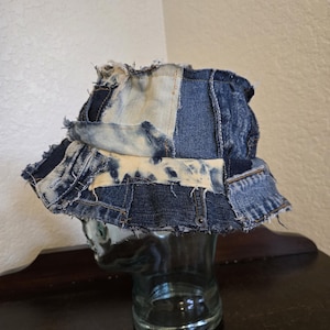 May include: A bucket hat constructed from patchwork denim fabric in various shades of blue and bleached white. The hat features a frayed, raw-edge design with visible seams, stitching, and a wide, downward-sloping brim made of multiple denim panels.