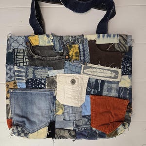 May include: A patchwork tote bag made from repurposed denim. The bag features a variety of blue denim patches, pockets, and a dark blue denim handle. The design includes a white pocket and an orange corduroy pocket.