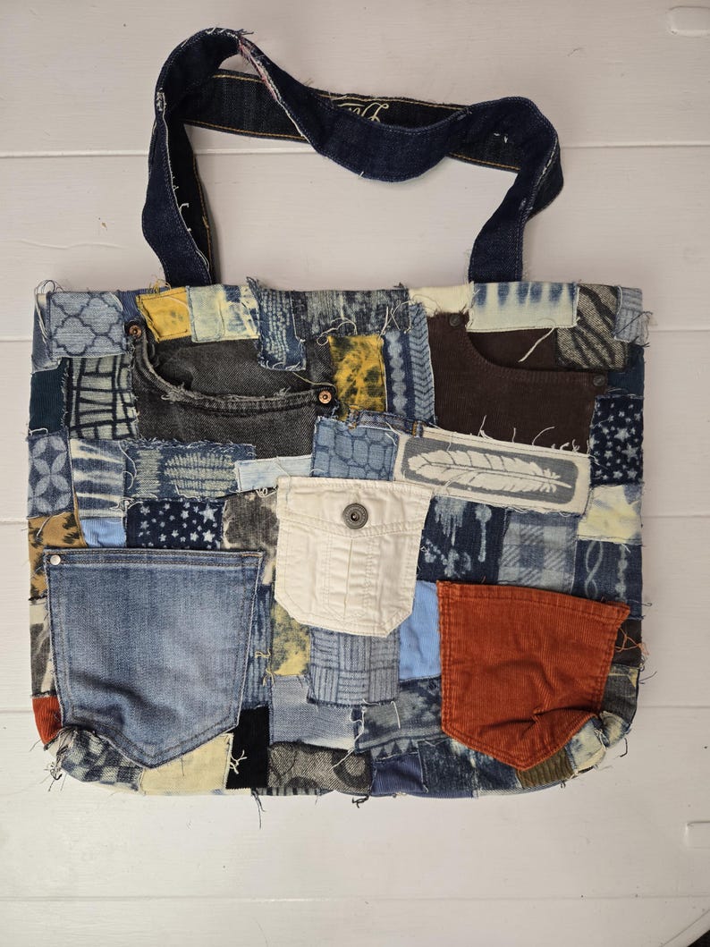 May include: A patchwork tote bag made from repurposed denim. The bag features a variety of blue denim patches, pockets, and a dark blue denim handle. The design includes a white pocket and an orange corduroy pocket.