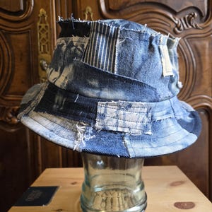 May include: A bucket hat constructed from various patches of blue denim fabric. The design features a mix of light and dark wash denim, including sections with vertical white stripes and frayed edges, creating a textured, layered appearance.