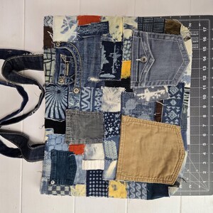 May include: A patchwork tote bag made from repurposed denim and other fabrics. The bag features a variety of blue denim patches, pockets, and decorative stitching. The bag has a dark blue denim handle and a rectangular shape.