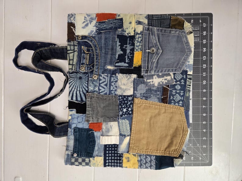 May include: A patchwork tote bag made from repurposed denim and other fabrics. The bag features a variety of blue denim patches, pockets, and decorative stitching. The bag has a dark blue denim handle and a rectangular shape.