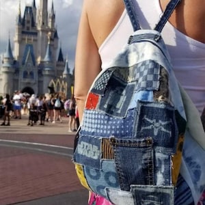 Peut inclure: Un sac à dos patchwork en denim avec des bretelles réglables. Le sac à dos présente divers patchs en denim, dont un logo Star Wars et d'autres motifs. L'arrière-plan montre un château et une foule de personnes.