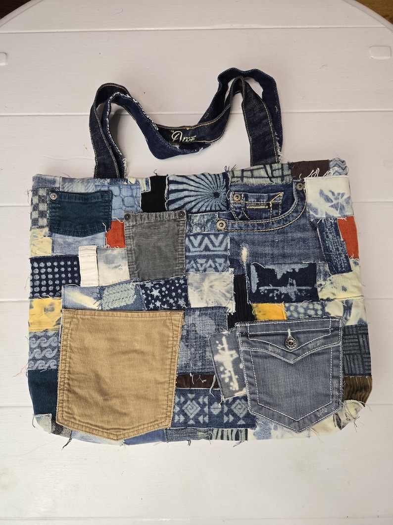 May include: A patchwork tote bag made from recycled denim and other fabrics. The bag features a variety of blue denim patches, corduroy pockets, and decorative stitching. The handles are made of denim. The word "Ame" is visible on the handle.