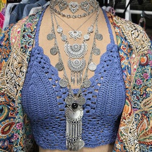 Puede incluir: Un top halter de ganchillo azul combinado con un collar plateado de varias capas con motivos de corazón, un colgante de loto, medallones semicirculares ornamentados y dijes de monedas colgantes, sobre un cárdigan abierto con estampado de cachemira.