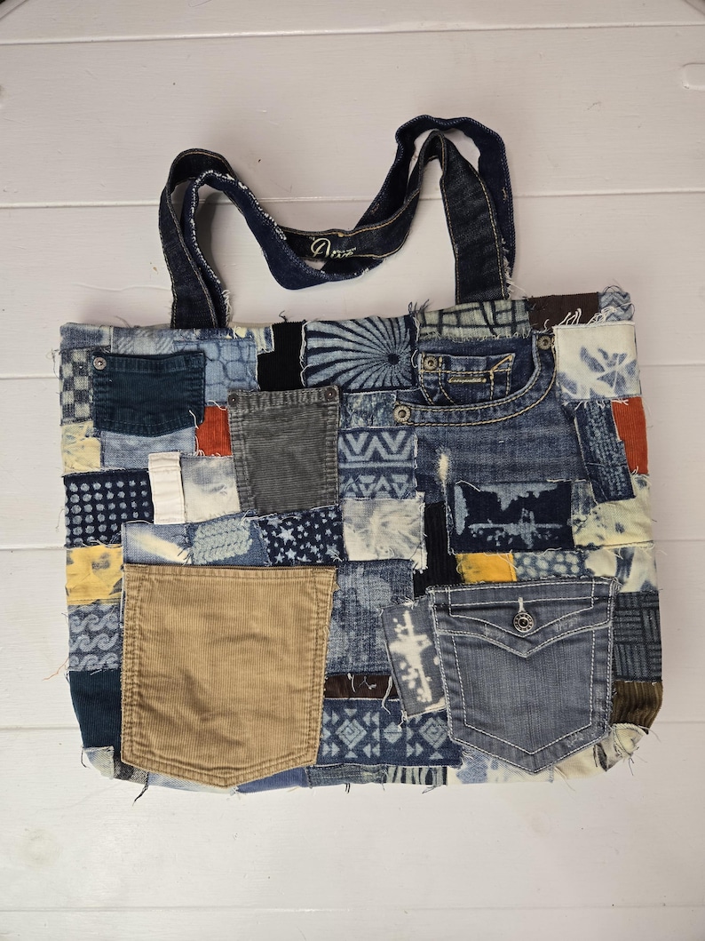 May include: A patchwork tote bag made from various denim pieces, featuring different shades of blue, brown, and tan. The bag includes pockets and a corduroy pocket. The handles are made of denim. The bag is on a white wooden surface.