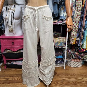 May include: Beige wide-leg pants featuring an elastic waistband with a drawstring, two front pockets with ruffled edges, and three horizontal rows of ruffled trim near the hemline. The pants have a loose, flowing silhouette and a light neutral color.