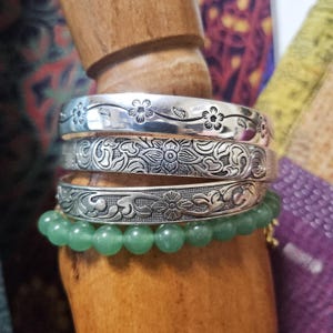 Puede incluir: Un conjunto de tres brazaletes de metal plateado con patrones grabados de flores y enredaderas, situados sobre una hilera de cuentas de piedra preciosa de color verde claro, lisas y redondas. La joyería se exhibe en un soporte de madera.