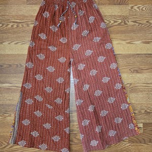 May include: Rust-colored wide-leg pants with a repeating floral pattern and vertical stripes. The pants have a drawstring waist and decorative trim along the side seams. The fabric appears to be a lightweight material.