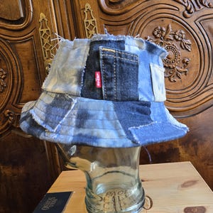 May include: A patchwork bucket hat constructed from various shades of blue denim fabric. The hat features frayed edges, visible stitching, and a small red Levi's brand tag on the side. The design incorporates multiple denim panels.
