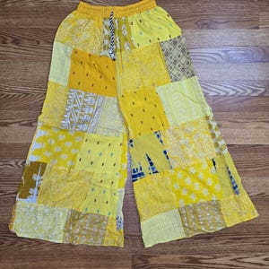May include: Yellow patchwork trousers with a drawstring waist. The trousers display a variety of yellow fabrics with different patterns, including floral and geometric designs. The trousers are wide-legged and appear to be made of a lightweight material.