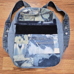 Puede incluir: Una mochila confeccionada con varios parches de tela de mezclilla azul. El diseño presenta un panel horizontal de pana negra, una cremallera azul oscuro y múltiples bolsillos de mezclilla. Las correas están hechas de tela de mezclilla reutilizada.