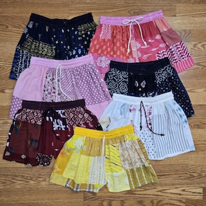 Op de afbeelding: Een verzameling patchwork shorts in verschillende kleuren en patronen. De shorts hebben elastische taillebanden met trekkoorden. Kleuren zijn onder meer roze, marineblauw, geel, rood en wit. De shorts zijn gemaakt van verschillende stoffen en ontwerpen.