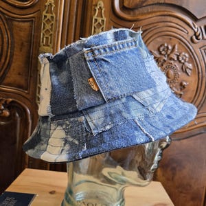 May include: A patchwork bucket hat constructed from various shades of blue denim fabric, featuring frayed edges, visible stitching, and a small orange rectangular tag on a side pocket detail. The hat is displayed on a clear glass head form.
