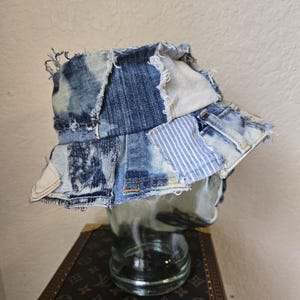 May include: A patchwork denim bucket hat. The hat is constructed from various shades of blue denim, including light wash, dark wash, and striped fabrics. The edges of the hat are frayed, giving it a distressed look. The hat is displayed on a glass head.