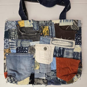 May include: A patchwork tote bag made from recycled denim. The bag features a variety of denim washes and textures, with pockets and a dark blue handle. The bag is rectangular and sits on a white surface.