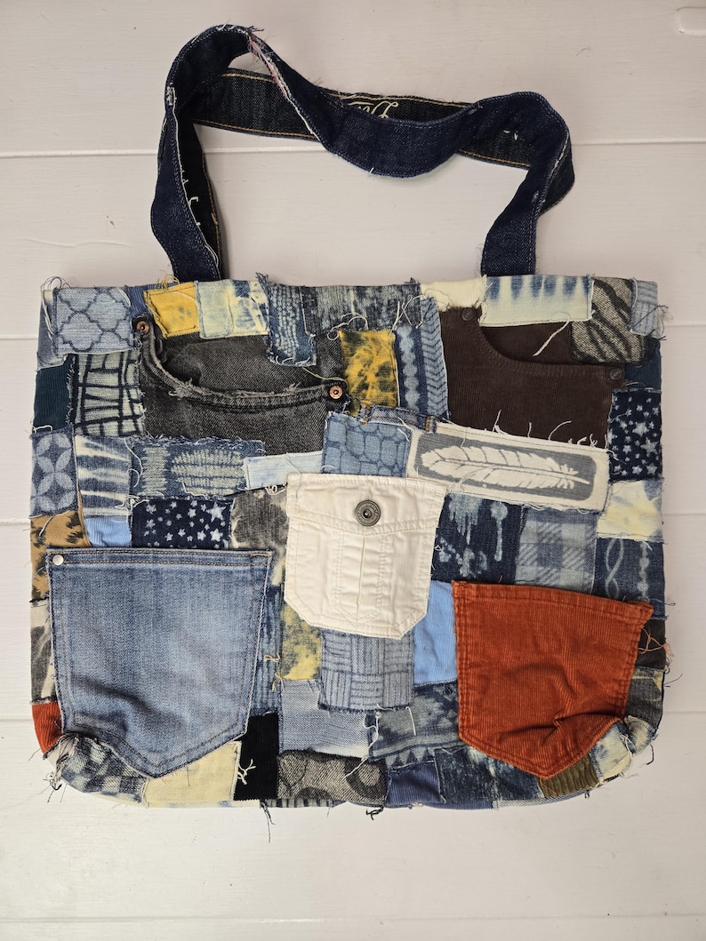 May include: A patchwork tote bag made from recycled denim. The bag features a variety of denim washes and textures, with pockets and a dark blue handle. The bag is rectangular and sits on a white surface.