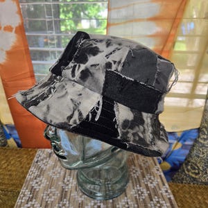 以下が含まれることがあります： 白黒のタイダイ柄のパッチワークバケットハット。帽子は縁がほつれており、ガラス製の頭部マネキンに飾られています。帽子は様々な生地で作られています。