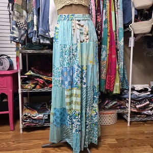 Peut inclure: Une jupe longue patchwork dans les tons de bleu, turquoise et blanc, présentée sur un mannequin. La jupe présente une variété de motifs floraux et géométriques. Un haut en crochet beige est associé à la jupe. La jupe est longue.