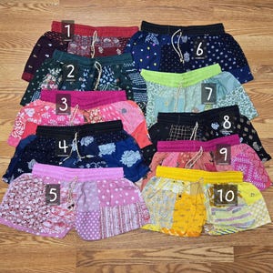 May include: A collection of ten pairs of colorful, patterned shorts. The shorts feature various designs, including floral, geometric, and patchwork patterns. Each pair has a drawstring waistband. The shorts are arranged on a wooden surface.