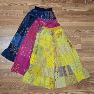 May include: Three long, patchwork skirts in shades of yellow, pink, and navy blue. Each skirt features a unique pattern of squares and rectangles, with a drawstring waistband. The skirts are made of a lightweight fabric.