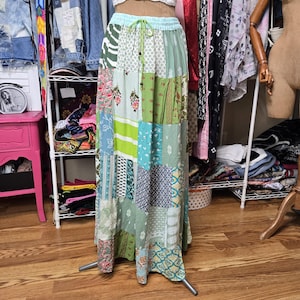 May include: A floor-length patchwork skirt featuring a variety of green, white, and blue fabric panels with floral, geometric, and striped patterns. The skirt has an elasticized waistband with a thin green drawstring and a loose, flowing fit.