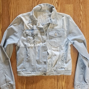 May include: Pale blue denim jacket with pinstriped sleeves and front panels. The jacket features two chest pockets and two side pockets. It is buttoned up and has a collar.
