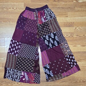 May include: Patchwork trousers in a wide-leg style, featuring a mix of purple, burgundy, and black panels. The design incorporates floral, geometric, and skull patterns. The waistband is a solid purple. The trousers are made from a lightweight material.