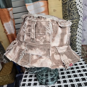 May include: A brown and white tie-dye bucket hat with a distressed, patchwork design. The hat features a frayed brim and a small red tag. The hat is made of denim material.