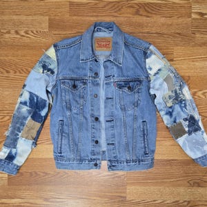 Könnte beinhalten: Hellblaue Jeansjacke mit Patchworkärmeln. Die Jacke hat einen klassischen Kragen, eine Knopfleiste und Taschen. Die Ärmel sind mit Flicken aus blauen und beigen Stoffen verziert, was einen einzigartigen, Used-Look erzeugt. Maße in cm.