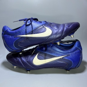 May include: Pair of blue and purple soccer cleats with white Nike swoosh logos. The cleats have metal studs on the soles and blue laces. The shoes are made of a shiny, textured material.