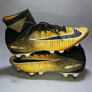 May include: Pair of black and yellow soccer cleats with the word "MERCURIAL" printed on the side. The cleats have a high-top design with black laces and a white Nike swoosh. The cleats are made of a synthetic material.