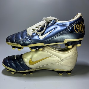 Könnte beinhalten: Paar Vintage Nike Fußballschuhe. Ein Schuh ist marineblau mit einem weißen Swoosh und der Zahl "90" in Gold. Der andere ist cremefarben mit einem goldenen Swoosh. Beide haben gelbe Stollen.