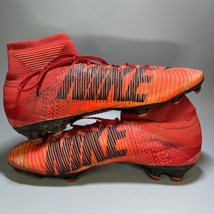 May include: Pair of red and orange Nike football boots. The boots have black accents and the Nike logo in black. The boots have a high-top design with laces and a textured pattern. The boots are designed for use on a football pitch.
