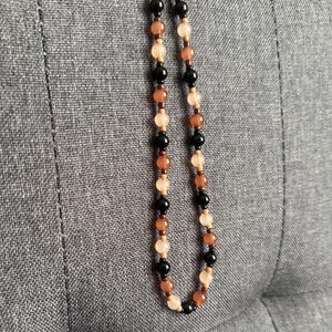 May include: A beaded necklace with a variety of earth-toned beads. The necklace features a mix of black, brown, peach, and cream-colored beads strung together. The necklace is hanging against a gray textured background.