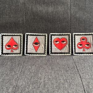 May include: Four square ceramic coasters with playing card suit designs: spade, diamond, heart, and club. Each coaster features a red suit symbol with eyes, set against a white and black patterned background, framed by a black border.