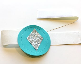 May include: A turquoise circle with a silver glitter diamond shape in the center. The circle is attached to a white strap with hook and loop fasteners.