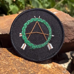 May include: A round black patch featuring a green wreath design with a brown letter "A" in the center. Two white arrows are positioned on either side of the wreath. The patch is displayed on a brown rock.