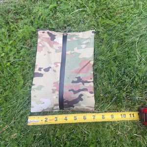 May include: A rectangular pouch featuring a multi-colored camouflage pattern in shades of green, brown, and tan. A vertical black zipper runs down the center. A yellow measuring tape placed along the bottom shows the pouch is 9 inches wide.