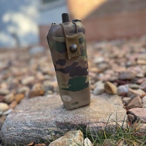 May include: A camouflage-patterned canteen with a black cap and a tan strap secured by a metal snap. The canteen is resting on a gray rock, with a blurred background of other rocks and foliage.